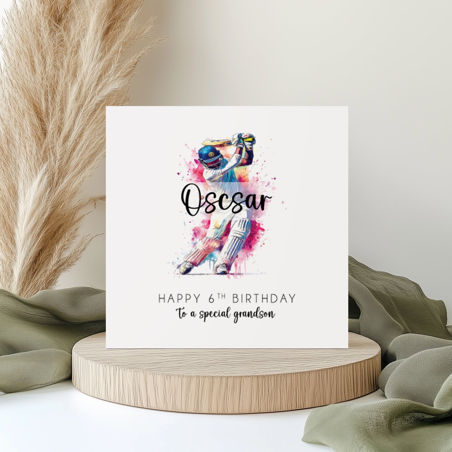 A square white birthday card featuring a vibrant watercolour illustration of a cricketer in full whites and blue helmet mid-batting stroke, surrounded by pink and blue paint splashes. The name "Oscar" is overlaid in bold cursive script. Text reads "Happy 6th Birthday to a special grandson" in spaced capitals and flowing script. Displayed on a wooden disc with sage green linen and pampas grass.