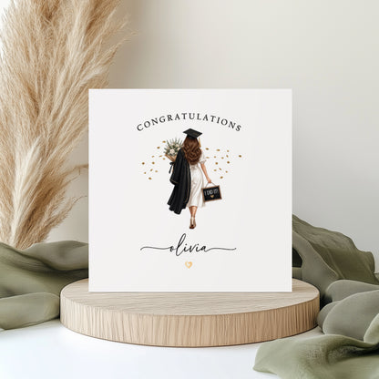 A square white graduation card featuring an illustration of a young woman viewed from behind, wearing a white dress and black graduation cap and gown. She is carrying a floral bouquet over one shoulder and a small chalkboard sign reading "I Did It!" in the other hand, surrounded by scattered gold confetti. The word "Congratulations" is printed in spaced capitals at the top of the card, with the name "Olivia" written in flowing cursive script below the illustration, accompanied by a small gold heart. The car
