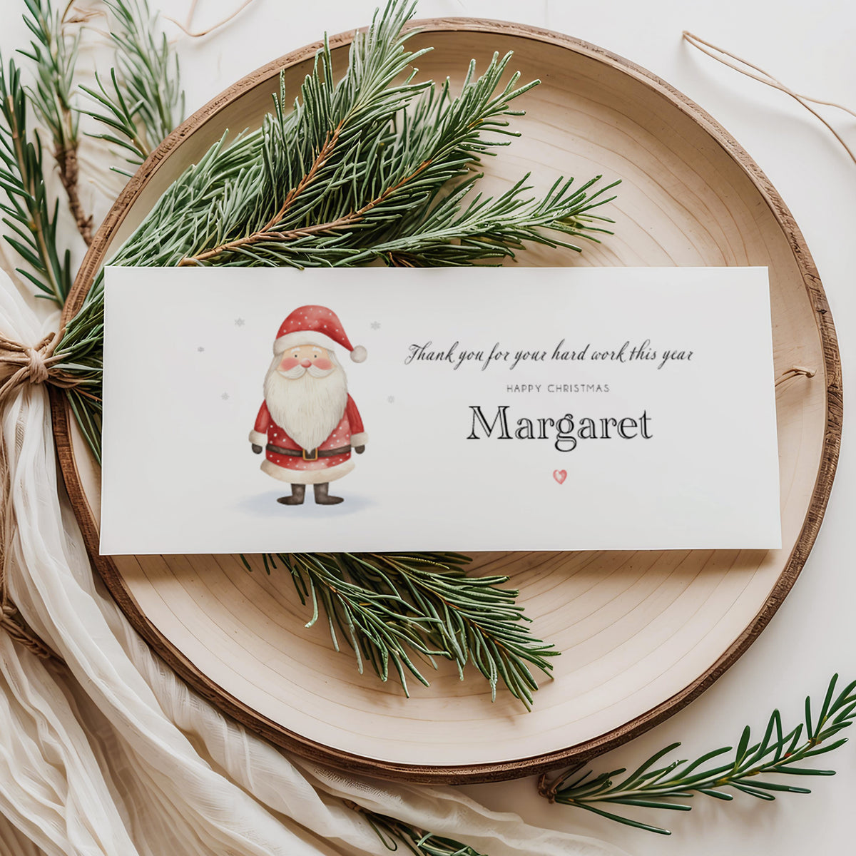 A rectangular white money wallet displayed flat on a wooden disc, featuring a watercolour illustration of a jolly Santa in red suit and boots on the left. To the right the text reads "Thank you for your hard work this year" in cursive script, "Happy Christmas" in small spaced capitals and "Margaret" in large elegant serif font, finished with a small red heart. Styled with pine sprigs and cream linen ribbon.