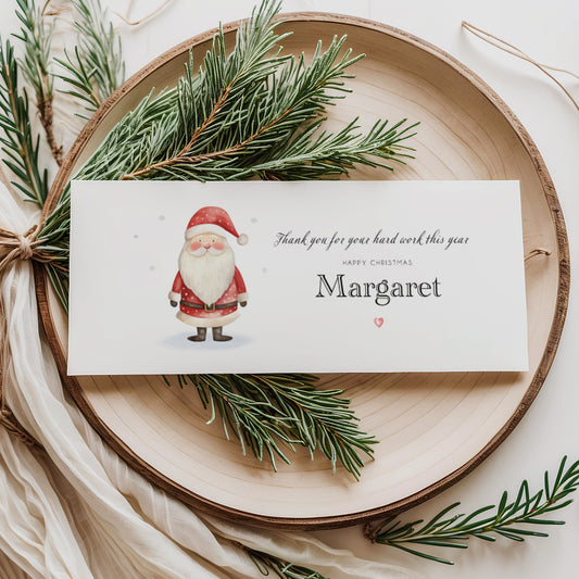 A rectangular white money wallet displayed flat on a wooden disc, featuring a watercolour illustration of a jolly Santa in red suit and boots on the left. To the right the text reads "Thank you for your hard work this year" in cursive script, "Happy Christmas" in small spaced capitals and "Margaret" in large elegant serif font, finished with a small red heart. Styled with pine sprigs and cream linen ribbon.