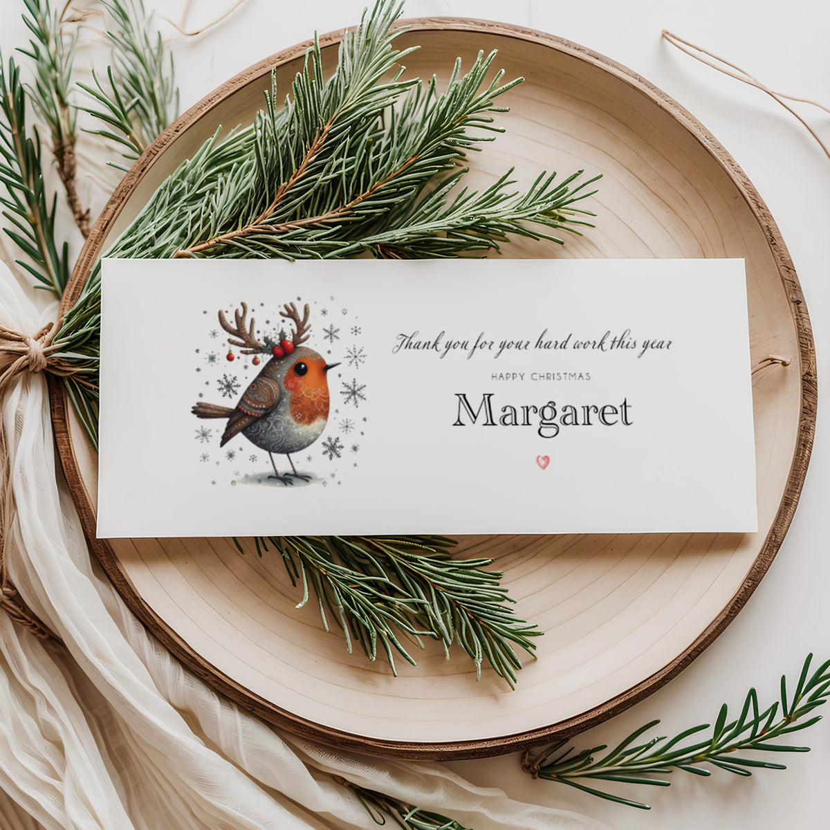 A rectangular white Christmas bonus money wallet displayed flat on a wooden disc, featuring an illustrated robin wearing festive reindeer antlers with red berries, surrounded by snowflake details. To the right the text reads "Thank you for your hard work this year" in cursive script, "Happy Christmas" in small spaced capitals and "Margaret" in large elegant serif font, finished with a small red heart. Styled with pine sprigs and cream linen ribbon.