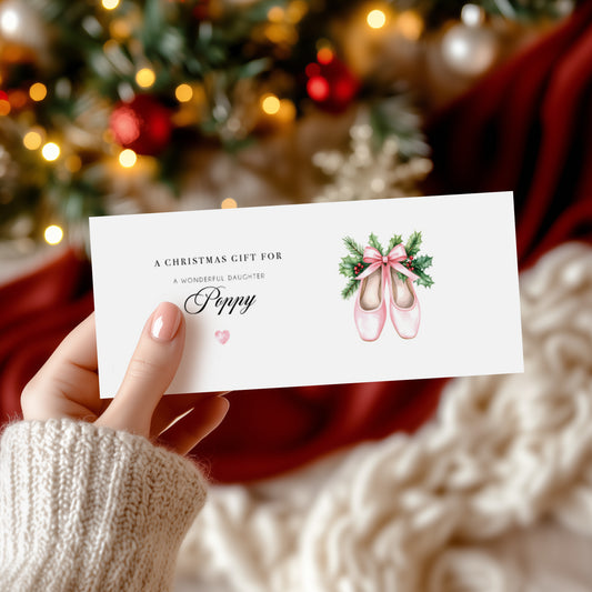 Hand holding a personalised Christmas gift money wallet card with ballet shoes illustration in front of a decorated tree.
