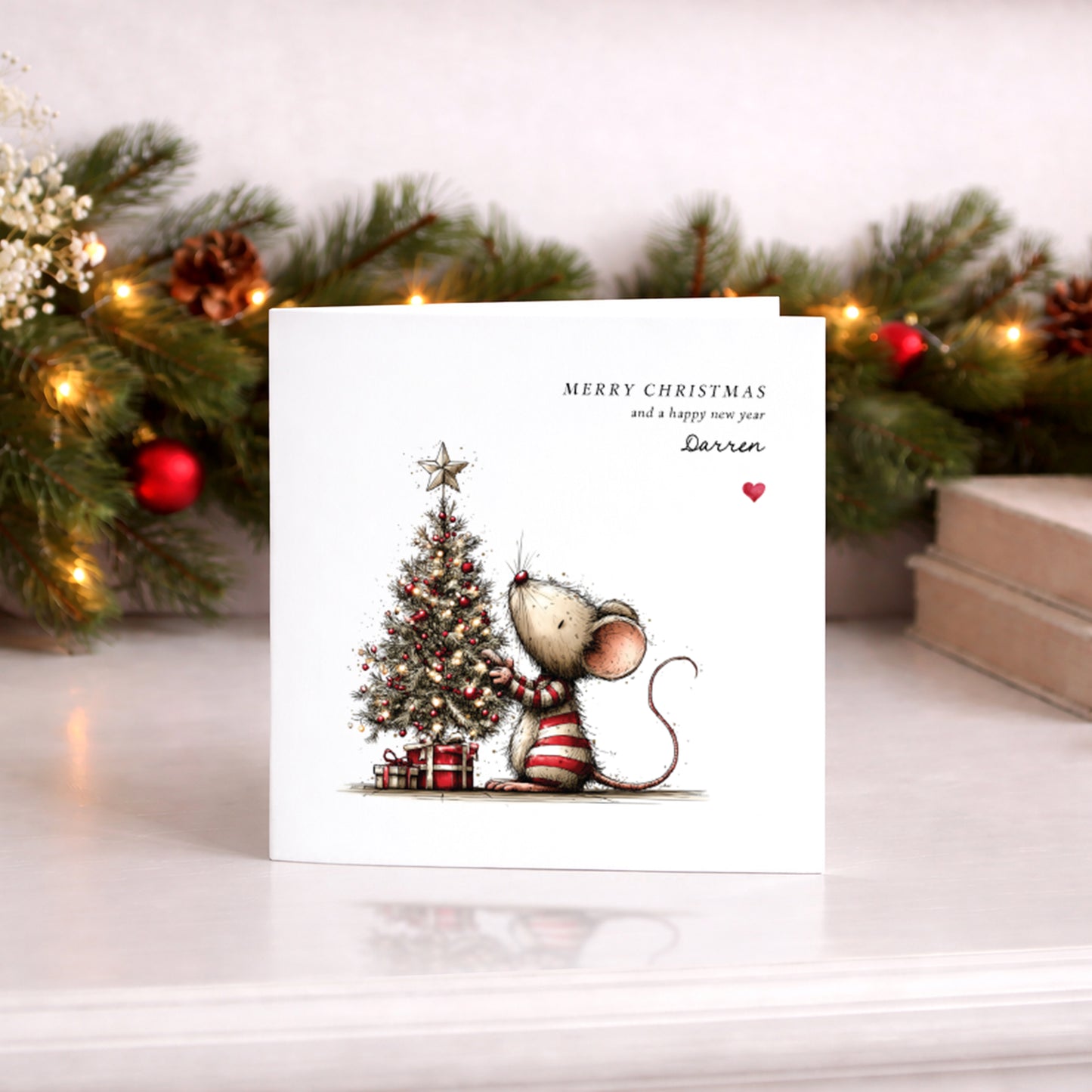 A square white Christmas card featuring an illustration of a small mouse in a red and white striped jumper decorating a glowing Christmas tree adorned with red baubles, fairy lights and a star on top, with wrapped gifts beneath. Text reads "Merry Christmas and a happy new year" with "Darren" in cursive script and a small red heart. Displayed against a lit pine garland with red baubles.
