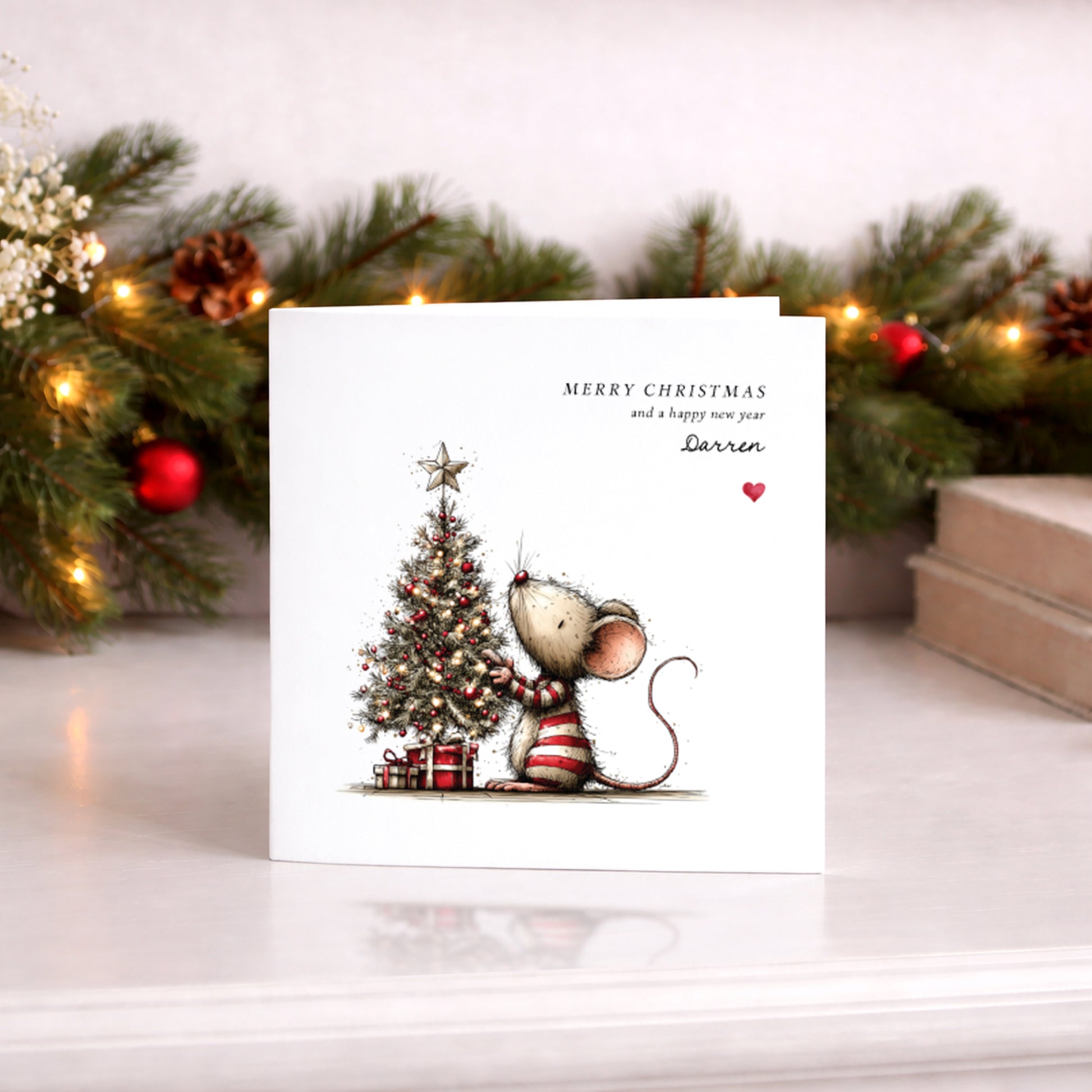 A square white Christmas card featuring an illustration of a small mouse in a red and white striped jumper decorating a glowing Christmas tree adorned with red baubles, fairy lights and a star on top, with wrapped gifts beneath. Text reads "Merry Christmas and a happy new year" with "Darren" in cursive script and a small red heart. Displayed against a lit pine garland with red baubles.