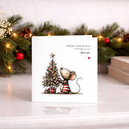 A square white Christmas card featuring an illustration of a small mouse in a red and white striped jumper decorating a glowing Christmas tree adorned with red baubles, fairy lights and a star on top, with wrapped gifts beneath. Text reads "Merry Christmas and a happy new year" with "Darren" in cursive script and a small red heart. Displayed against a lit pine garland with red baubles.