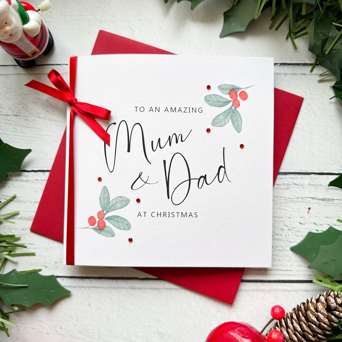 A square white Christmas card featuring watercolour holly berry sprigs with sage green leaves and red berries in the upper right and lower left corners, accented with small scattered red gem embellishments. The text reads "to an Amazing" in small spaced capitals at the top, with "Mum & Dad" in large flowing black cursive script in the centre, and "at Christmas" in small spaced capitals below. The card is tied with a real red satin ribbon bow on the left side and sits on a matching red envelope on a white pa