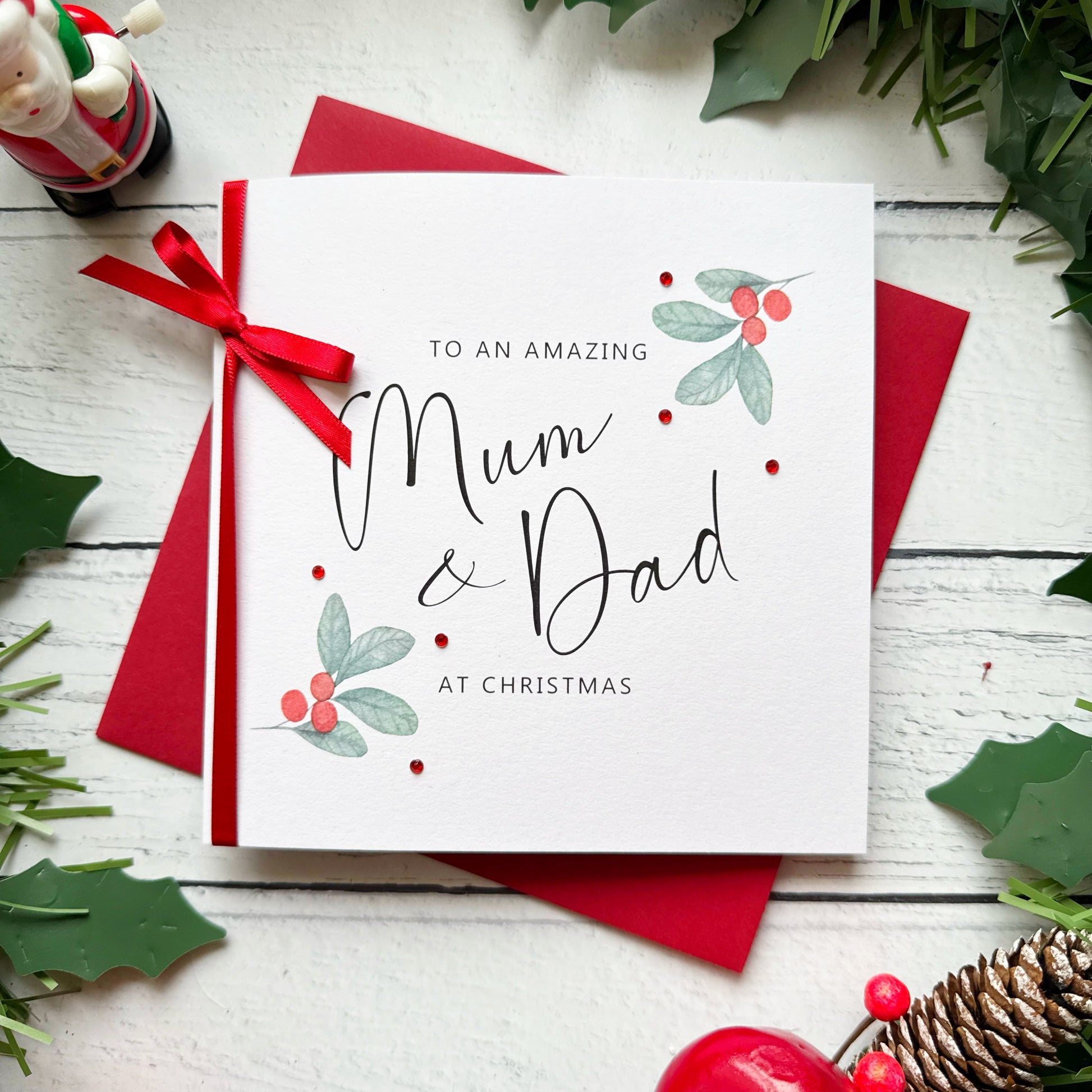 A square white Christmas card featuring watercolour holly berry sprigs with sage green leaves and red berries in the upper right and lower left corners, accented with small scattered red gem embellishments. The text reads "to an Amazing" in small spaced capitals at the top, with "Mum & Dad" in large flowing black cursive script in the centre, and "at Christmas" in small spaced capitals below. The card is tied with a real red satin ribbon bow on the left side and sits on a matching red envelope on a white pa