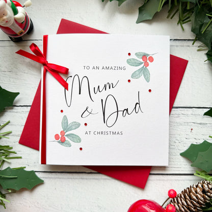 A square white Christmas card featuring watercolour holly berry sprigs with sage green leaves and red berries in the upper right and lower left corners, accented with small scattered red gem embellishments. The text reads "to an Amazing" in small spaced capitals at the top, with "Mum & Dad" in large flowing black cursive script in the centre, and "at Christmas" in small spaced capitals below. The card is tied with a real red satin ribbon bow on the left side and sits on a matching red envelope on a white pa
