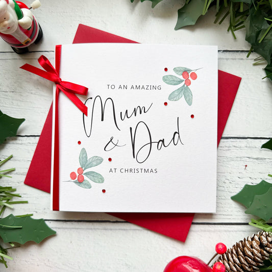 A square white Christmas card featuring watercolour holly berry sprigs with sage green leaves and red berries in the upper right and lower left corners, accented with small scattered red gem embellishments. The text reads "to an Amazing" in small spaced capitals at the top, with "Mum & Dad" in large flowing black cursive script in the centre, and "at Christmas" in small spaced capitals below. The card is tied with a real red satin ribbon bow on the left side and sits on a matching red envelope on a white pa