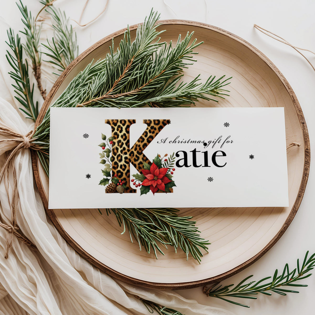 A rectangular white Christmas money wallet displayed flat on a wooden disc, featuring a large bold leopard print letter "K" decorated with a red poinsettia, holly leaves, red berries and pine cone details. The text "A Christmas gift for" sits above the name "Katie" in large elegant black serif font, with small snowflake details scattered around. Styled with pine sprigs and cream linen ribbon.
