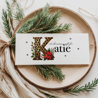 A rectangular white Christmas money wallet displayed flat on a wooden disc, featuring a large bold leopard print letter "K" decorated with a red poinsettia, holly leaves, red berries and pine cone details. The text "A Christmas gift for" sits above the name "Katie" in large elegant black serif font, with small snowflake details scattered around. Styled with pine sprigs and cream linen ribbon.