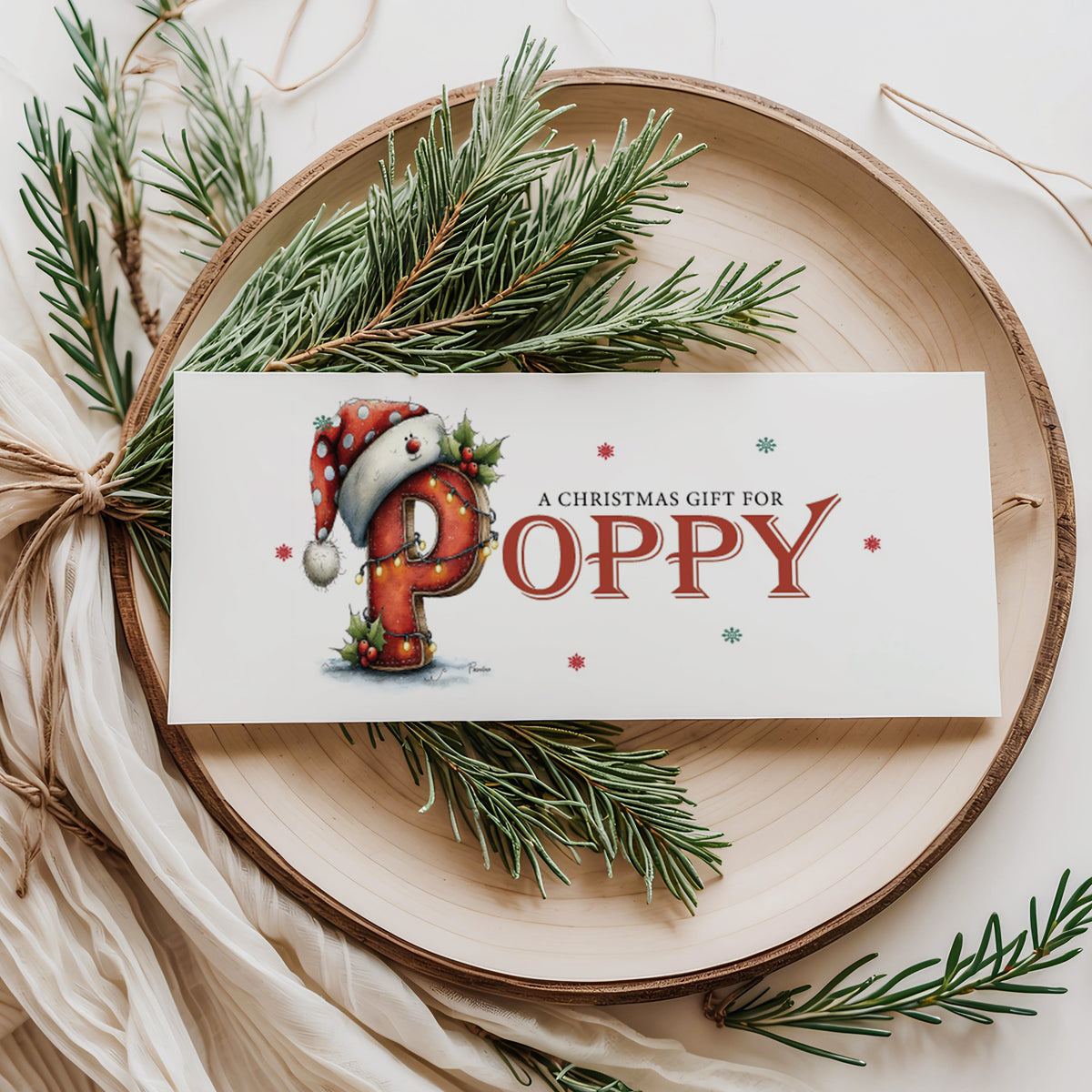 A rectangular white Christmas money wallet displayed flat on a wooden disc, featuring a large illustrated festive letter "P" wearing a Santa hat with polka dots, decorated with holly and fairy lights. The text "A Christmas Gift for" sits above the name "POPPY" in large bold red serif font, with red and green snowflake details scattered around. Styled with pine sprigs and cream linen