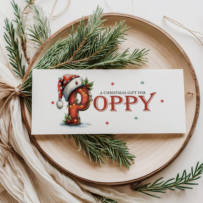 A rectangular white Christmas money wallet displayed flat on a wooden disc, featuring a large illustrated festive letter "P" wearing a Santa hat with polka dots, decorated with holly and fairy lights. The text "A Christmas Gift for" sits above the name "POPPY" in large bold red serif font, with red and green snowflake details scattered around. Styled with pine sprigs and cream linen