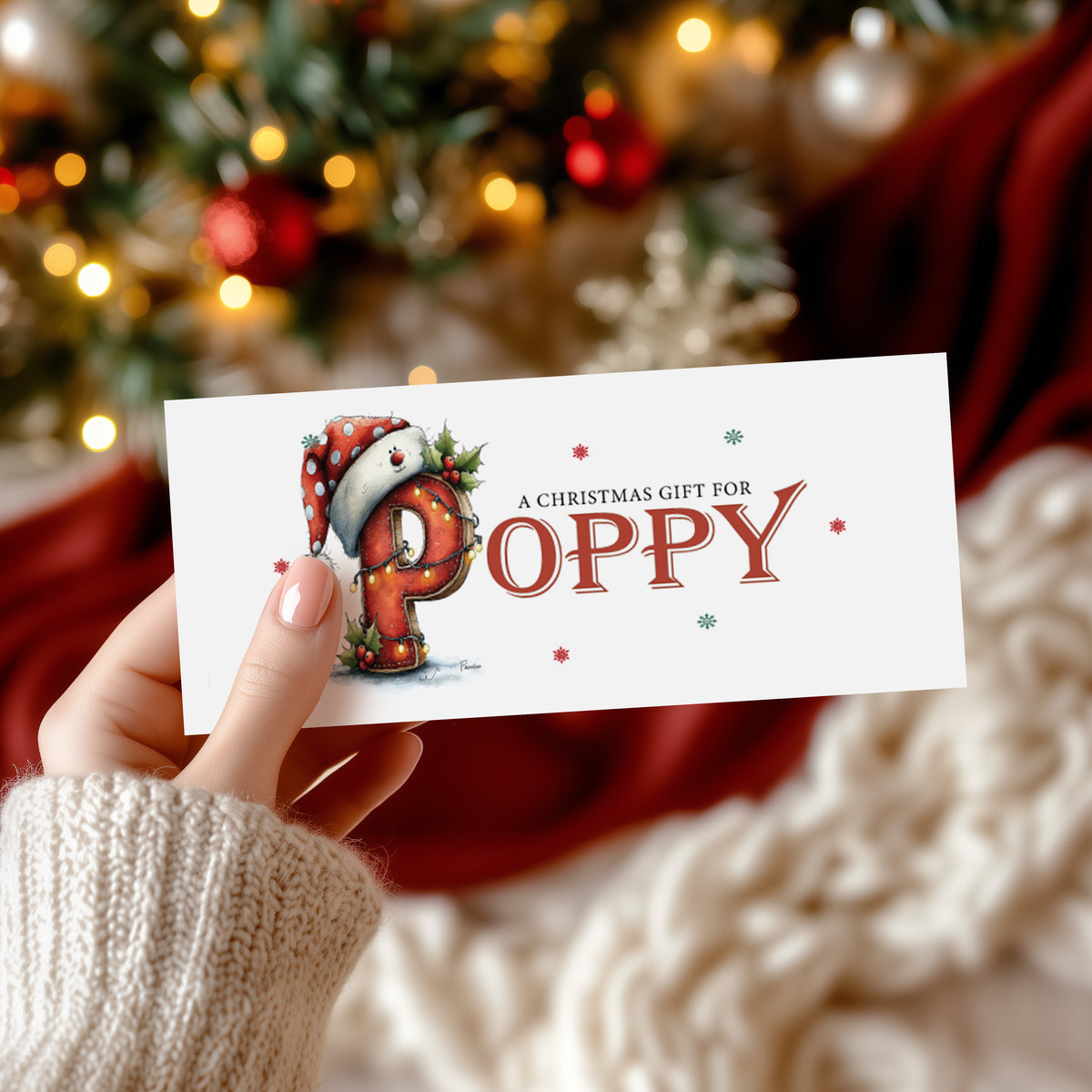 A rectangular white Christmas money wallet held in hand, featuring a large illustrated festive letter "P" wearing a polka dot Santa hat, decorated with holly and fairy lights. The text "A Christmas Gift for" sits above the name "POPPY" in large bold red serif font, with red and green snowflake details scattered around. Displayed against a blurred decorated Christmas tree with red and gold baubles.