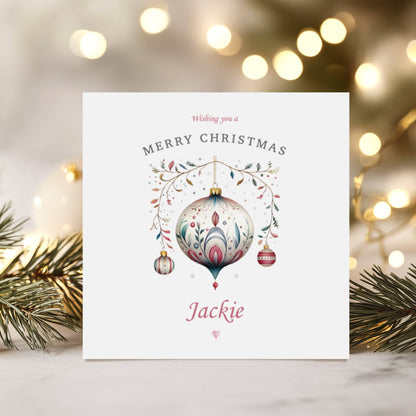 A square white Christmas card featuring a watercolour illustration of a large ornate vintage bauble in soft cream, pink and teal with intricate floral and botanical detailing, suspended from a delicate gold wire adorned with small leaves and berries in pink, teal and green. Two smaller baubles in teal and red hang either side. Above the illustration the text reads "Wishing you a" in small pink italics, followed by "Merry Christmas" in spaced grey capitals. Below, the name "Jackie" is printed in large pink c