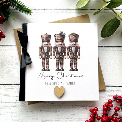 A square white Christmas card featuring an illustration of three leopard print nutcracker soldiers in rich bronze and copper tones, each wearing a decorated military cap, standing side by side. Below the illustration the text reads "Merry Christmas" in flowing cursive script, followed by "to a Special Family" in small spaced capitals, finished with a small wooden heart embellishment. The card is tied with a real black satin ribbon bow on the left side and sits on a matching kraft envelope on a white painted