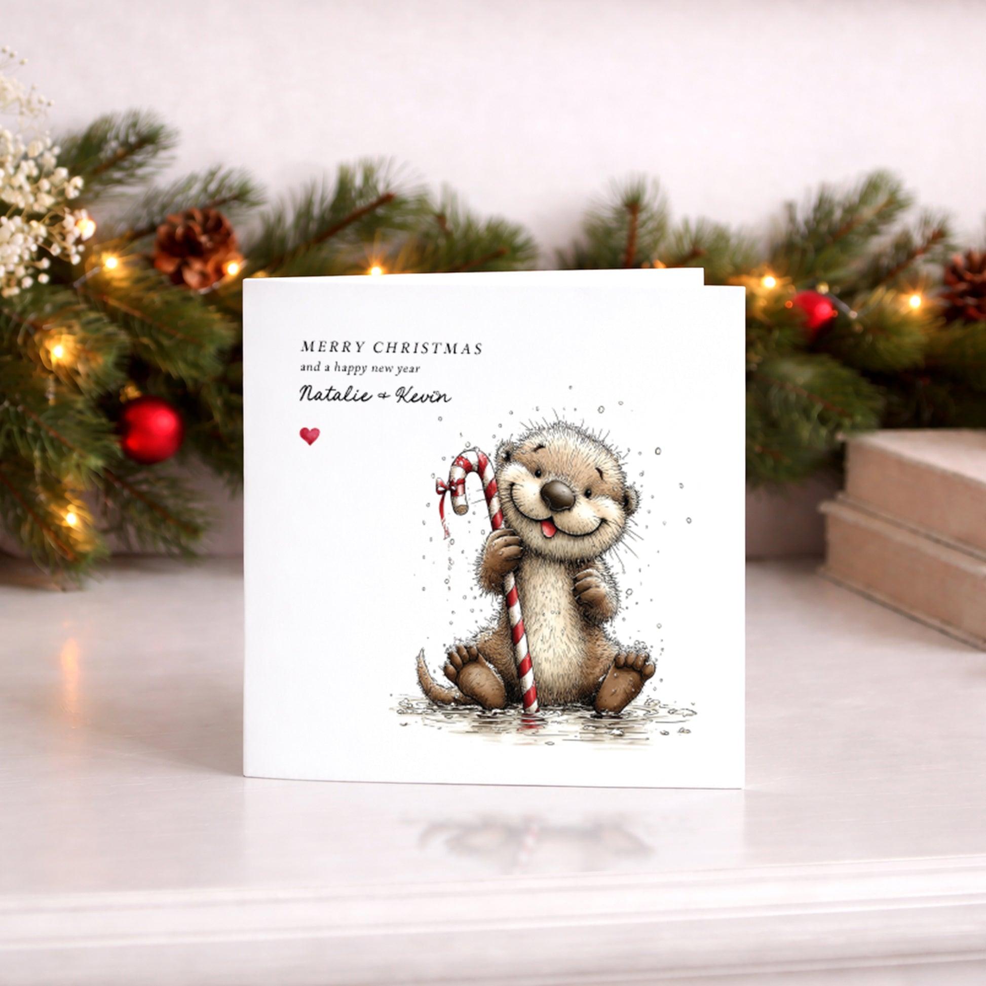 A square white Christmas card featuring an illustration of a fluffy smiling otter sitting in water holding a red and white candy cane, with snowflakes scattered around. Text reads "Merry Christmas and a happy new year" with "Natalie & Kevin" in cursive script and a small red heart. Displayed upright against a lit pine garland with red baubles and pine cones.