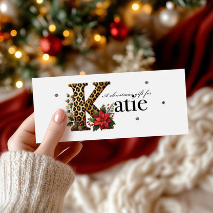 A rectangular white Christmas money wallet held in hand, featuring a large bold leopard print letter "K" decorated with a red poinsettia, holly leaves, red berries and pine cone details. The text "A Christmas gift for" sits in elegant cursive script above the name "Katie" in large black serif font, with small snowflake details scattered around. Displayed against a blurred decorated Christmas tree with red and gold baubles.