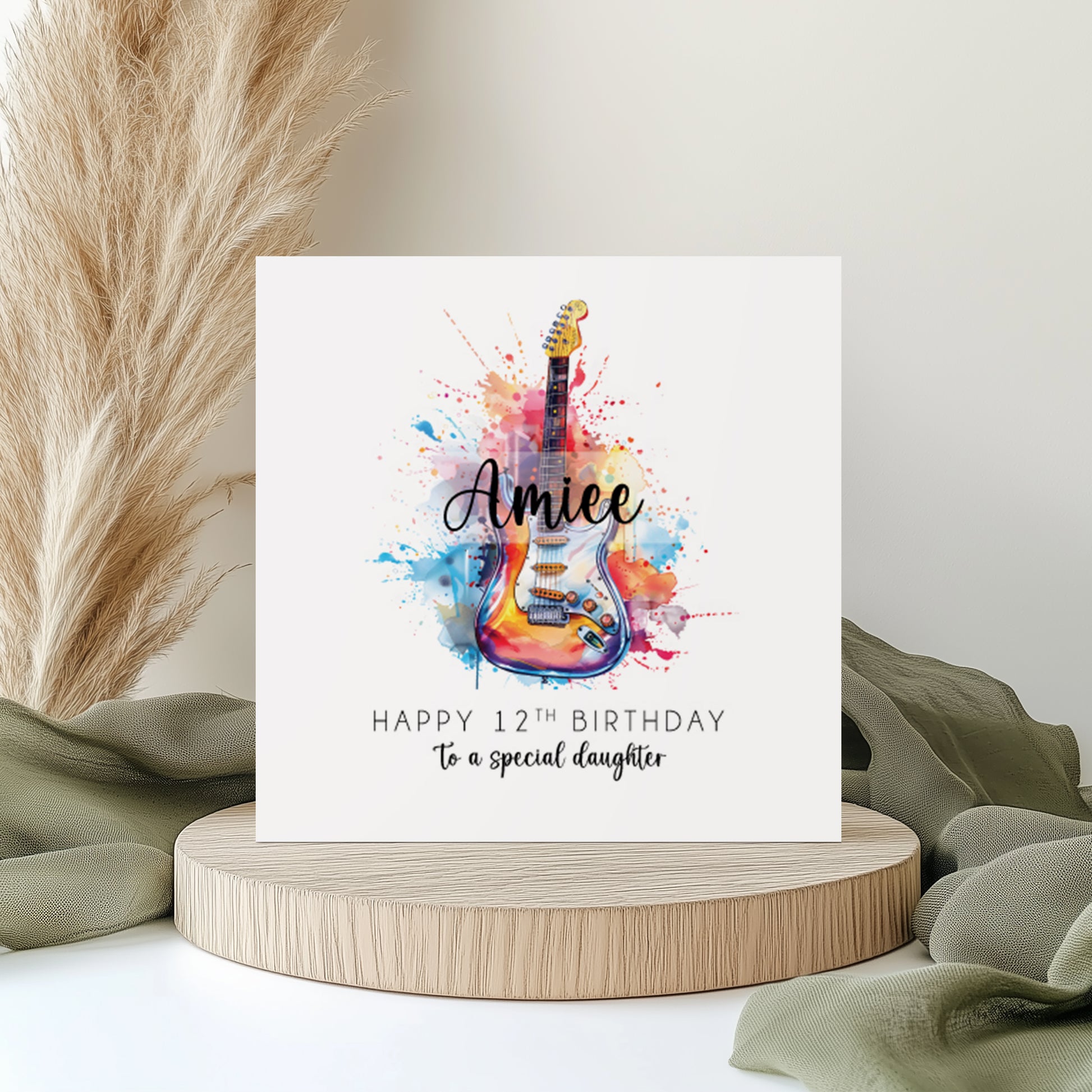 Personalised birthday card featuring a colourful watercolour-style electric guitar with vibrant paint splash effects. The card reads “Happy 12th Birthday to a special daughter” with the custom name “Amiee” across the guitar design. Displayed on a neutral styled background with pampas grass and soft fabric accents.