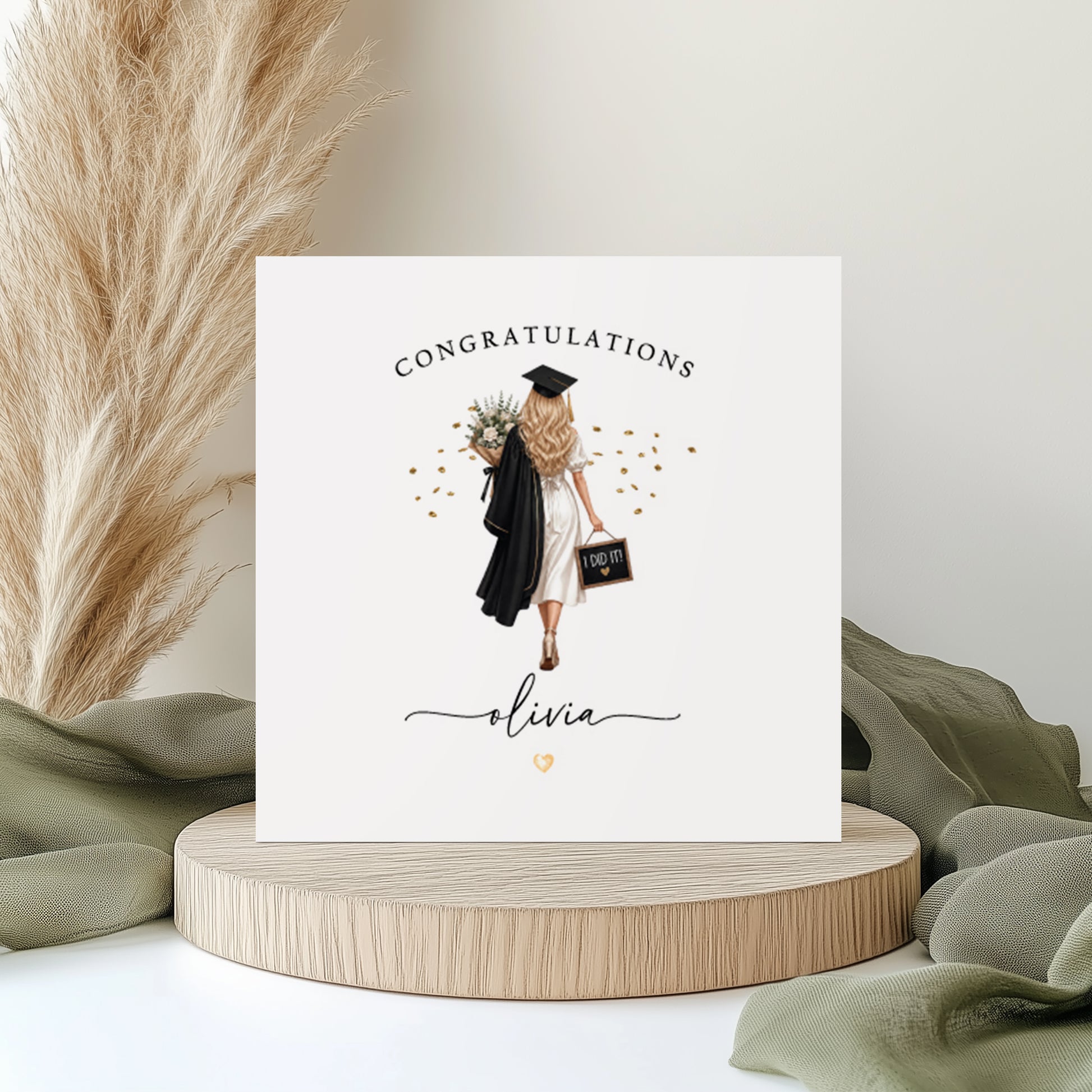 A square white graduation card featuring an illustration of a young woman viewed from behind, wearing a white dress and black graduation cap and gown. She is carrying a floral bouquet over one shoulder and a small chalkboard sign reading "I Did It!" in the other hand, surrounded by scattered gold confetti. The word "Congratulations" is printed in spaced capitals at the top of the card, with the name "Olivia" written in flowing cursive script below the illustration, accompanied by a small gold heart. The car