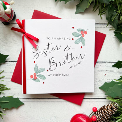 A square white Christmas card featuring watercolour holly berry sprigs with sage green leaves and red berries in the upper right and lower left corners, accented with small scattered red gem embellishments. The text reads "to an Amazing" in small spaced capitals at the top, with "Sister & Brother-in-Law" in large flowing black cursive script in the centre, and "at Christmas" in small spaced capitals below. The card is tied with a real red satin ribbon bow on the left side and sits on a matching red envelope