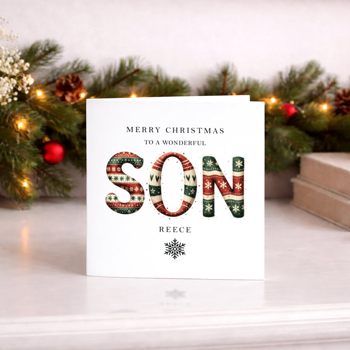 A square white Christmas card featuring large bold letters spelling "SON" in a festive knitted jumper style pattern of red, green and cream with snowflakes, stars and heart details. Text reads "Merry Christmas to a Wonderful" in small spaced capitals above, with the name "Reece" in spaced capitals and a decorative snowflake below. Displayed upright against a lit pine garland with red baubles and pine cones.