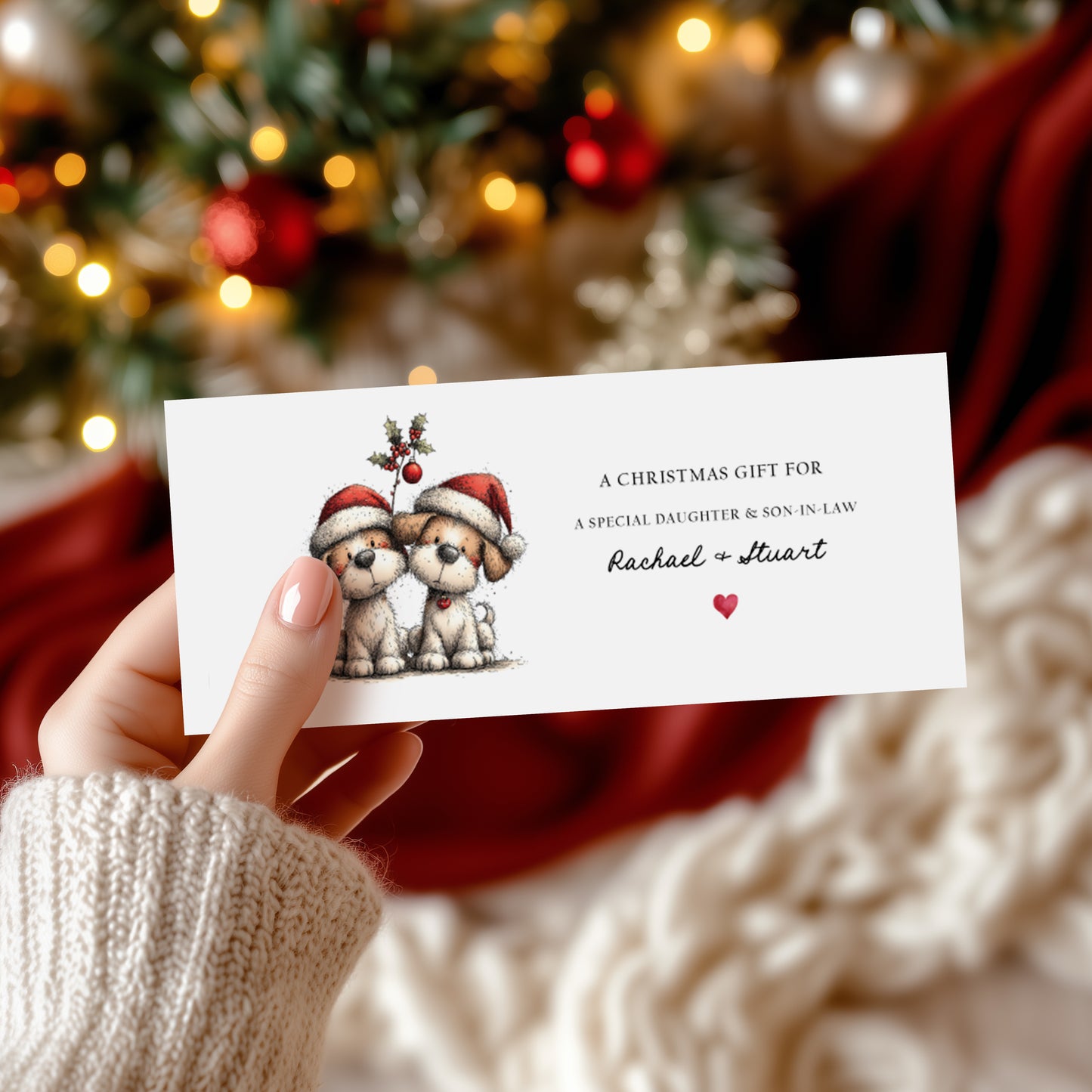 A rectangular white Christmas money wallet held in hand, featuring two fluffy dogs in red Santa hats beneath a holly sprig. Text reads "A Christmas Gift for a Special Daughter & Son-in-Law" with "Rachael & Stuart" in cursive script and a small red heart. Displayed against a blurred Christmas tree background with red and gold baubles.
