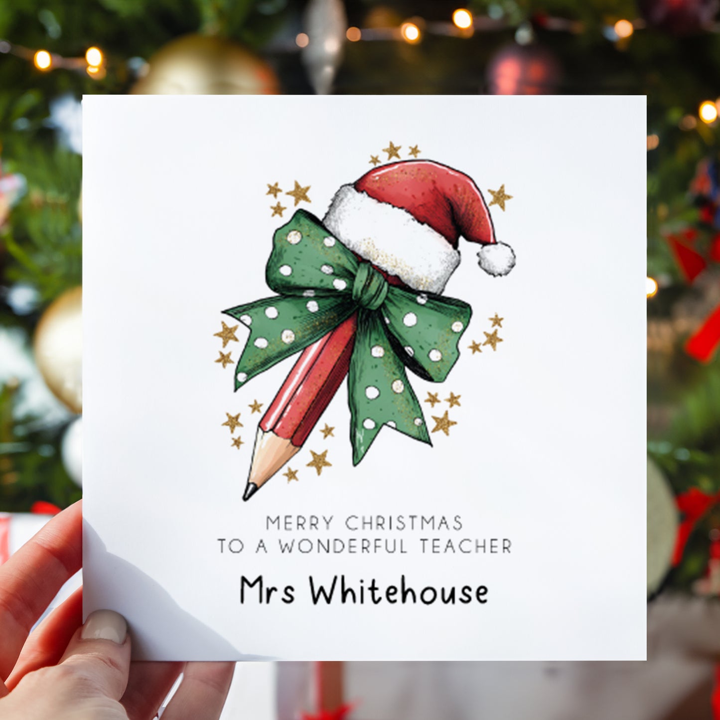 A hand holds a white personalized Christmas greeting card featuring an illustration of a red pencil wrapped in a green polka-dot bow with a Santa hat, with the text 'MERRY CHRISTMAS TO A WONDERFUL TEACHER Mrs Whitehouse'. The background shows a blurred Christmas tree with lights and ornaments.