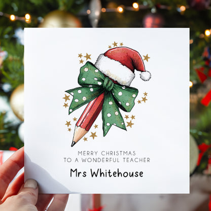 A hand holds a white personalized Christmas greeting card featuring an illustration of a red pencil wrapped in a green polka-dot bow with a Santa hat, with the text 'MERRY CHRISTMAS TO A WONDERFUL TEACHER Mrs Whitehouse'. The background shows a blurred Christmas tree with lights and ornaments.