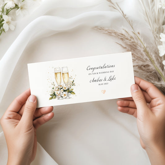 A rectangular white money wallet featuring a watercolour illustration of two champagne flutes surrounded by white blossom flowers, green leaves and gold confetti dots. To the right of the illustration the text reads "Congratulations on Your Wedding Day" with "Ambao & Luke" in cursive script and the date "01.07.2027" below, finished with a small peach heart. The wallet is displayed flat on a white linen cloth, styled alongside a kraft gift box with a cream ribbon bow, eucalyptus stems, dried pampas grass and