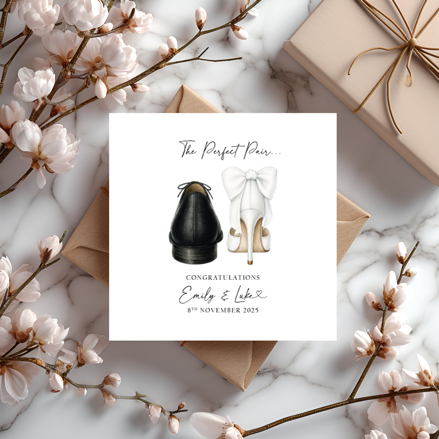 A square white wedding card featuring an illustration of two shoes viewed from behind — a black leather brogue and a white stiletto heel with a large white bow detail — sitting side by side. Above the illustration the text reads "The Perfect Pair..." in flowing cursive script. Below, "Congratulations" is printed in spaced capitals, followed by "Emily & Luke" in cursive script with a small heart, and the date "8th November 2025" beneath. The card is displayed on a kraft envelope on a white marble surface