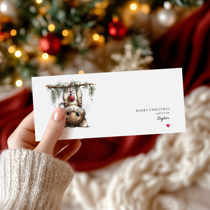 A hand in a cream knit sweater holds a white Christmas greeting card featuring a watercolor illustration of a cat hanging upside down from a pine branch with red ornaments. The card has the text 'MERRY CHRISTMAS a gift for you Taylor' and is photographed against a festive blurred background of a Christmas tree and red fabric.
