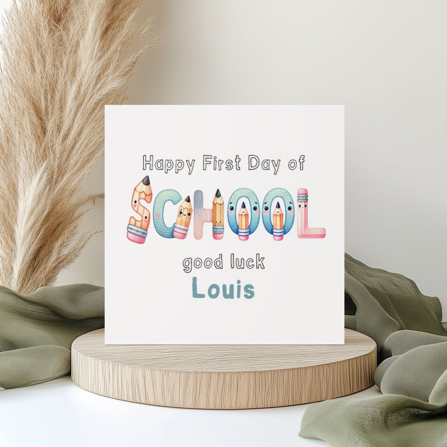 Personalised first day of school greeting card featuring colourful school-themed lettering made from pencils and stationery characters. The card reads “Happy First Day of School, good luck Louis” in playful pastel colours. Displayed on a neutral styled background with pampas grass and soft fabric accents.