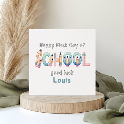 Personalised first day of school greeting card featuring colourful school-themed lettering made from pencils and stationery characters. The card reads “Happy First Day of School, good luck Louis” in playful pastel colours. Displayed on a neutral styled background with pampas grass and soft fabric accents.