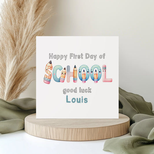 Personalised first day of school greeting card featuring colourful school-themed lettering made from pencils and stationery characters. The card reads “Happy First Day of School, good luck Louis” in playful pastel colours. Displayed on a neutral styled background with pampas grass and soft fabric accents.