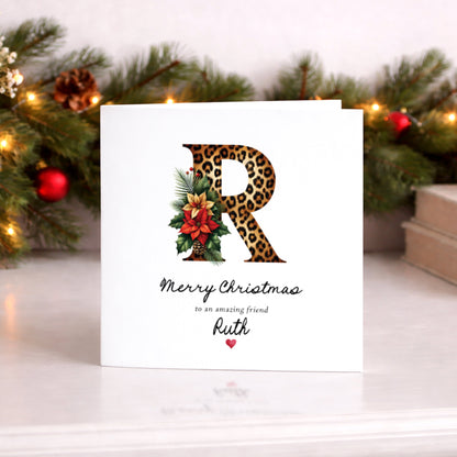 A square white Christmas card featuring a large bold leopard print letter "R" decorated with a festive arrangement of red poinsettia, holly, pine sprigs and red berries. Text reads "Merry Christmas" in flowing cursive script, "to an amazing friend" in small italics and "Ruth" in large cursive script below, finished with a small red heart. Displayed upright on a white surface against a lit pine garland with red baubles and candles.